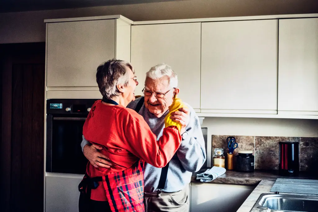 Dos personas mayores sonrientes abrazadas bailando la cocina de su casa