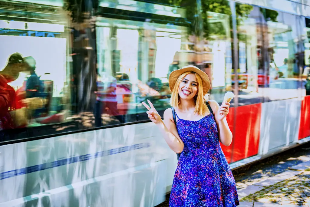Mujer sonriente con vestido azul y bus detras, con un helado en una mano y haciendo la uve con la otra